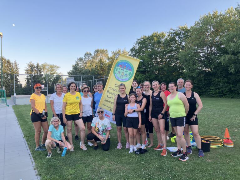 Die Teilnehmerinnen und Teilnehmer von Bewegt im Park stehen für ein Gruppenfoto neben der gelben Projektfahne auf der Wiese.