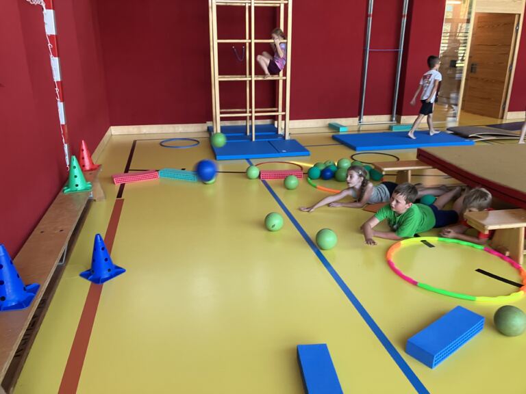 Kinder bewegen sich in einer Bewegungslandschaft aus Reifen, Bällen, Bänken und Sprossenwand im Turnsaal.