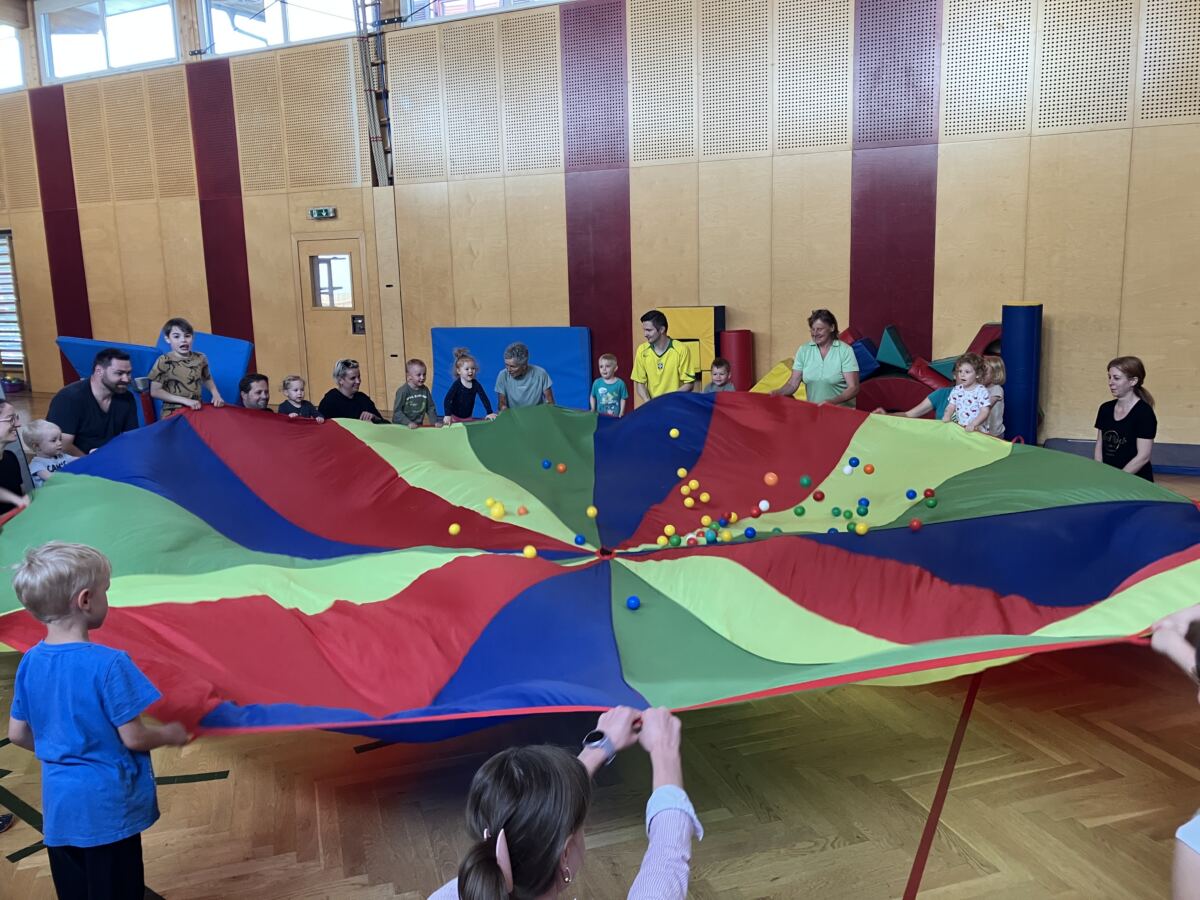 Eltern und Kleinkinder halten gemeinsam ein großes buntes Schwungtuch, auf dem bunte Bälle springen.