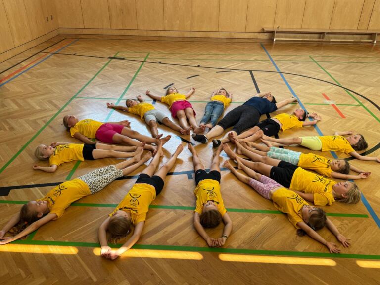 Kinder in gelben Sportcamp-Shirts liegen im Kreis auf dem Hallenboden, die Füße zur Mitte, und entspannen sich.