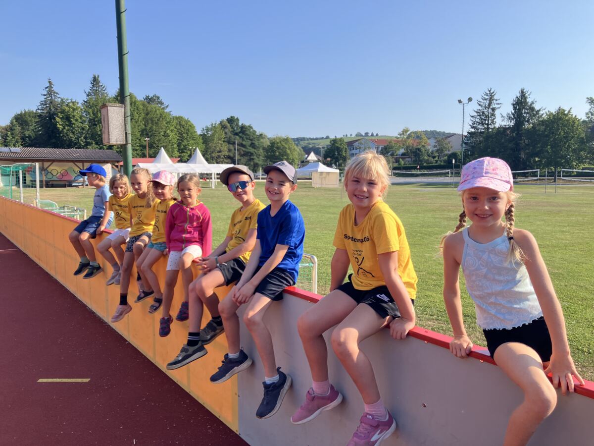 Neun Kinder in gelben Sportcamp-Shirts sitzen nebeneinander auf der Bande des Funcourts, dahinter der Sportplatz.